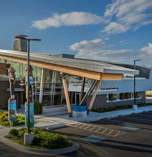 Modern building with a unique angled roof, featuring totem poles and a parking lot under a cloudy sky.