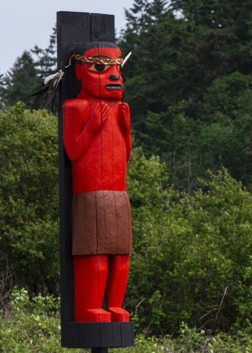 Carved Red Figure Black Post Wears Brown Skirt A carved red figure on a black post wears a brown skirt and a feathered headdress.