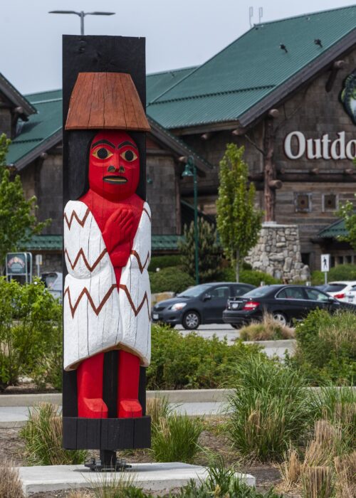 Red White Carved Wooden Totem Pole Stands Large A red and white carved wooden totem pole stands in front of a large building with a green roof.