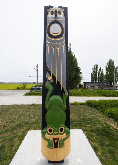 Totem Pole Carved Owl Top Frog Bottom Set A totem pole carved with an owl at the top and a frog at the bottom, set against a rural landscape.